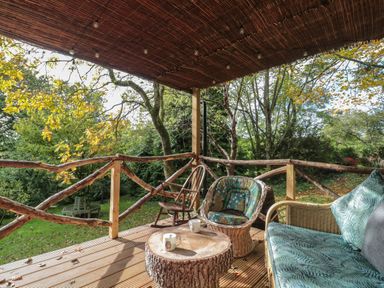A wooden deck with wicker and wooden chairs a round tree stump table and a garden view at Pear Tree Lodge in Combe near Langport