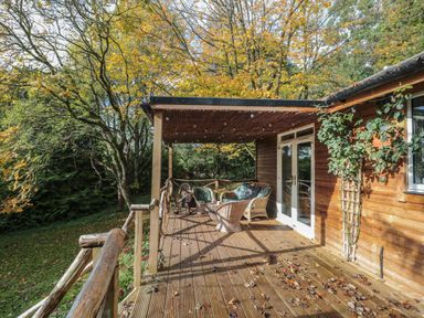 A wooden deck with outdoor furniture and a covered roof outside a wooden house surrounded by trees at Pear Tree Lodge in Combe near Langport