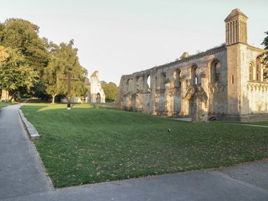 A stone ruined building with arched windows near a grassy area with a wooden cross and a paved path