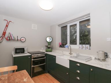 A kitchen with dark green cabinets a white farmhouse sink a black oven a microwave a kettle and a window above the sink at Pear Tree Lodge in Combe near Langport