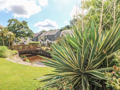 A garden with shrubs near a wooden bridge over water and houses in the background at Manuka in Falmouth