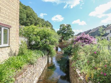 A narrow stream flowing between stone walls with plants and flowers alongside houses and a wooden footbridge in the background at Manuka in Falmouth