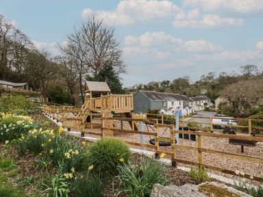 A fenced playground with wooden play structures and swings next to flowering plants and houses at Manuka in Falmouth