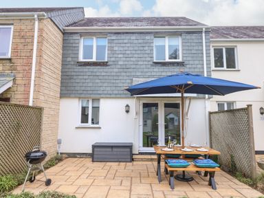 A patio with a wooden picnic table and benches under a blue umbrella a barbecue grill and a storage box outside a two-story house at Manuka in Falmouth
