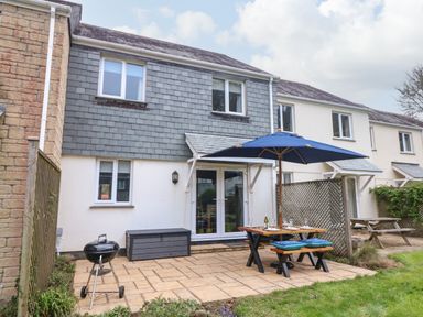 A backyard patio with a black grill a wooden picnic table with blue umbrella and cushions outside a two story house at Manuka in Falmouth