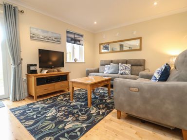 A living room with grey sofas a wooden coffee table and TV stand a patterned blue rug and wall art at Manuka in Falmouth