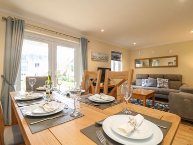 A dining table set with plates glasses and napkins next to a living area with sofas a coffee table and wall decorations at Manuka in Falmouth