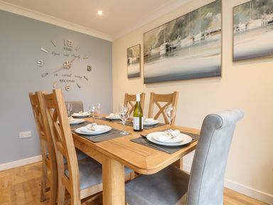 A dining room with a wooden table set for six with plates wine glasses and placemats and wall clock and paintings at Manuka in Falmouth