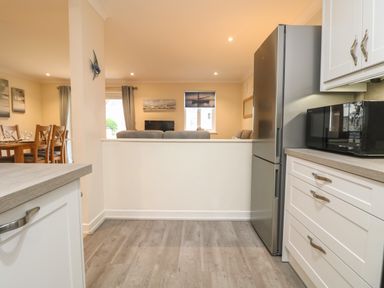 A kitchen area with white cabinets a microwave and a refrigerator overlooking a dining area with a wooden table and chairs at Manuka in Falmouth