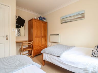 A bedroom with two single beds a wooden wardrobe a small desk with a chair a wall-mounted TV and a beach painting at Manuka in Falmouth