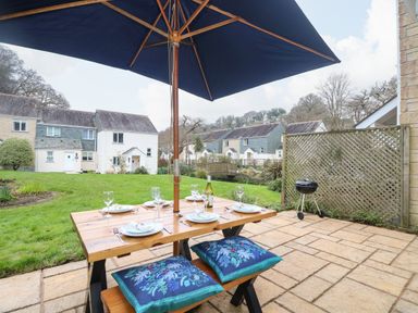 An outdoor patio with a wooden table set for four under a large umbrella with houses and green lawn in the background at Manuka in Falmouth