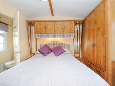A bedroom with a double bed with purple pillows wooden cabinets above and beside the bed and a wardrobe in Little Malory in St Giles-On-The-Heath