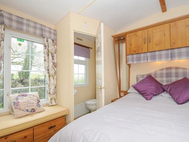 A bedroom with a bed with purple pillows and wooden cabinets above a window seat with a cushion and a view into a small bathroom at Little Malory in St Giles-On-The-Heath