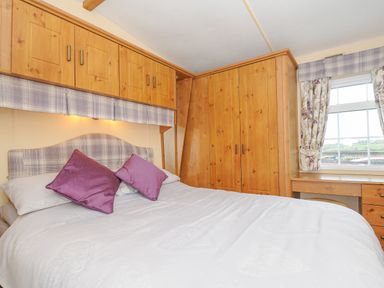 A bedroom with a double bed with purple pillows wooden overhead cabinets wooden wardrobes a window and a desk at Little Malory in St Giles-On-The-Heath