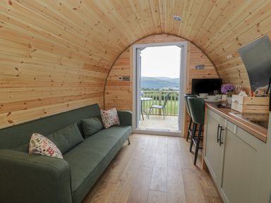 A pod interior with a green sofa, kitchen area with stools, and an open door to a deck with chairs at Tarrenhendre Pod in Llanegryn near Tywyn