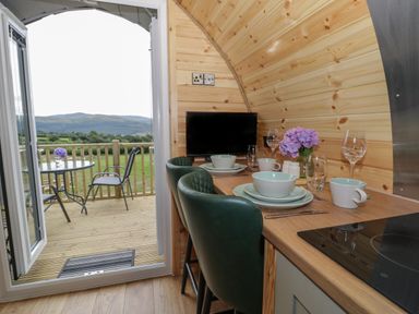 A small dining area with plates, bowls, cups, wine glasses, a television, and flowers on a wooden counter with green chairs and an open door leading to a balcony with a table and chairs in the countryside at Tarrenhendre Pod in Llanegryn near Tywyn