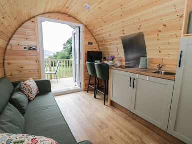 A small wooden pod interior with a green sofa pillows kitchen area with sink kettle two chairs and an open door to a deck with outdoor chair at Tarrenhendre Pod Llanegryn near Tywyn