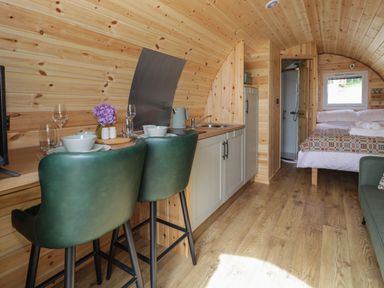 A small wooden pod interior with a kitchenette a dining area with two green chairs and a bed with a window at Tarrenhendre Pod in Llanegryn near Tywyn