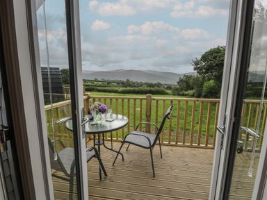 A wooden deck with a glass table and two chairs overlooking a green field and hills at Tarrenhendre Pod in Llanegryn near Tywyn