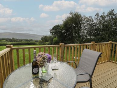 A wooden deck with a glass table holding a bottle, flowers, and two glasses next to a chair with fields and trees beyond at Tarrenhendre Pod in Llanegryn near Tywyn