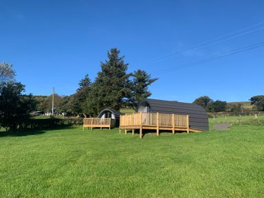 Two small cabins with wooden decks on grassy land with trees and clear blue sky at Tarrenhendre Pod in Llanegryn near Tywyn
