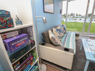 A living room with a white shelf of board games a beige sofa with patterned pillows a blue coffee table and large windows showing a green lawn at Culver View in Sandown