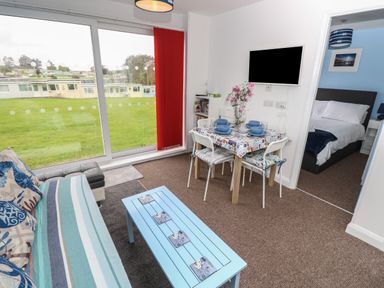 A living room with a striped sofa blue coffee table dining table set with blue dishes wall mounted tv and a bedroom visible at Culver View in Sandown
