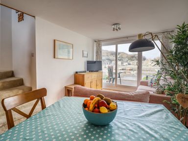 A dining table with a bowl of fruit in a living room with sofas a television and sliding glass doors overlooking a harbor at Branwen Porthmadog