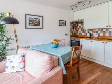 A kitchen with a dining table covered with a blue tablecloth and chairs next to a pink sofa with a cushion and a floor lamp in Branwen Porthmadog