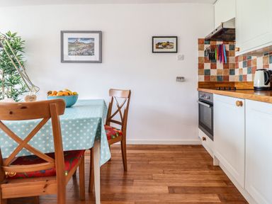 A kitchen and dining area with a table covered by a blue tablecloth with white flowers wooden chairs a bowl of fruit a potted plant tiled backsplash with knives and an oven at Branwen in Porthmadog