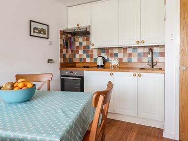 A kitchen with a wooden table covered with a blue patterned tablecloth and wooden chairs a bowl of fruit is on the table cabinets oven and sink are visible Branwen Porthmadog