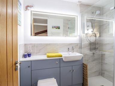 A bathroom with a sink on a blue vanity unit a large illuminated mirror and a glass shower enclosure at Branwen in Porthmadog