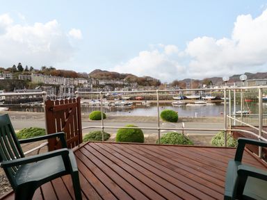 A wooden deck with plastic chairs overlooking a harbor with moored boats and buildings on hills at Branwen in Porthmadog