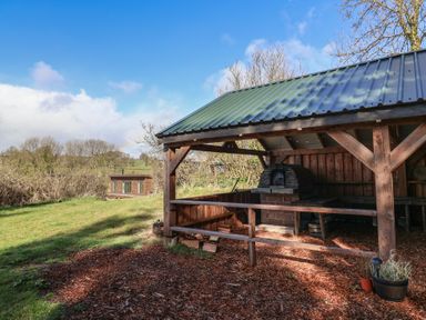 An outdoor covered wooden shelter with a brick oven and wood chips on the ground at Primrose in Higher Wambrook near Chard