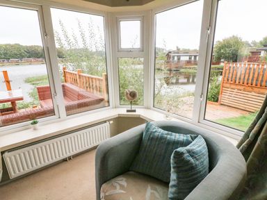 A corner seating area with a chair and cushions by large windows overlooking a deck and water at Lakeside 12 in Carnforth