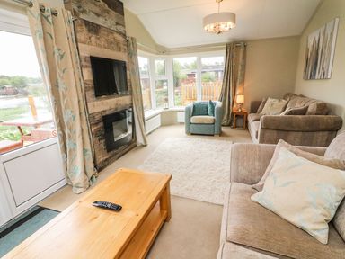 A living room with two beige sofas a blue armchair a wooden coffee table a wall-mounted tv and a fireplace at Lakeside 12 in Carnforth