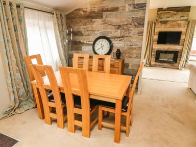 A dining area with wooden table and six chairs near floral curtains and a wall clock at Lakeside 12 in Carnforth