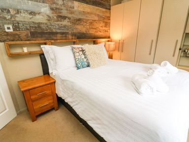 A bedroom with a double bed with pillows and rolled towels next to a wooden bedside table and wardrobe at Lakeside 12 in Carnforth