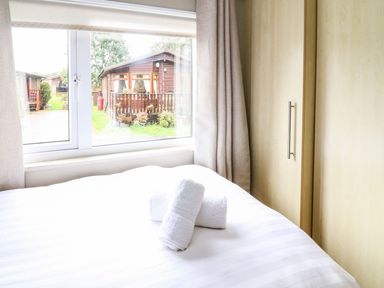 A bedroom with a bed with rolled towels near a window overlooking wooden cabins at Lakeside 12 in Carnforth