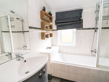 A bathroom with a white sink, a bathtub, wall-mounted wooden shelves, and a window with a dark blind at Lakeside 12 in Carnforth