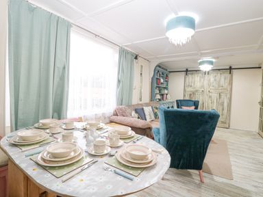 A dining table set with plates cups and utensils next to a living room with blue armchairs a sofa and a cabinet at White Rabbit Cross Lane in Eccles-On-Sea