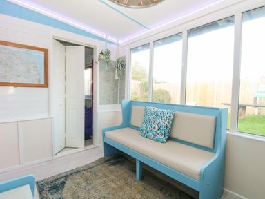 A sunroom with a blue bench and floral pillow beside windows showing a garden at White Rabbit Cross Lane in Eccles-On-Sea