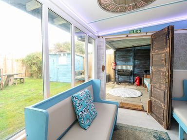 A seating area with blue and beige benches and a door opening to a covered patio with a barbecue and a blue shed in the garden at White Rabbit Cross Lane in Eccles-On-Sea