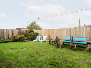 A grassy backyard with wooden fence garden beds two white lounge chairs and a long wooden table with chairs and bench at White Rabbit Cross Lane in Eccles-On-Sea
