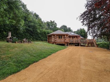 A log cabin with a wooden deck and stairs beside a dirt path with a grassy area and outdoor fireplace at Copper Lodge in Chulmleigh