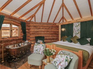 A bedroom with a wood log wall a bathtub near a window and floral cushions at Copper Lodge in Chulmleigh