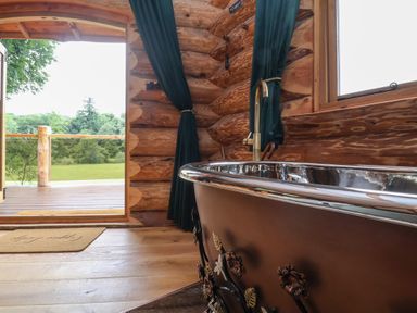 A wooden bathroom with a metal bathtub near a window and an open door showing a porch and grass outside at Copper Lodge in Chulmleigh