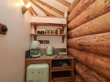 A small kitchen area with a teal SMEG coffee maker kettle and fridge next to wooden shelves and log wall at Copper Lodge in Chulmleigh