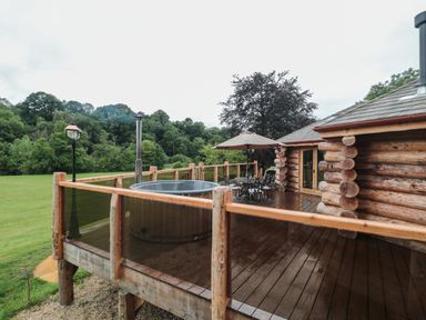 An outdoor wooden deck with a hot tub a table with chairs and an umbrella surrounded by trees at Copper Lodge in Chulmleigh