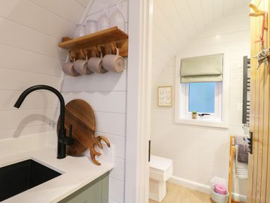 A small kitchen area with a black faucet and wooden mugs hanging above next to a bathroom with a toilet and a window with a green blind at Yes Deer in Ashbourne